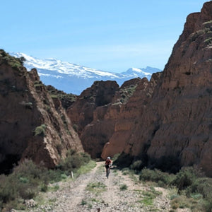Gravel rider cycling up a track through stunning red rocks, with the snow-capped Sierra Nevada mountains in the background, part of a scenic cycling route on Hidden Tracks Cycling's Flamenco Gravel trip