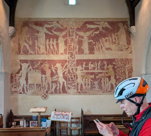 Medieval Ladder of Salvation wall painting in Chaldon church, Surrey, showing souls climbing a ladder out of purgatory past demons, with a gravel cyclist in a helmet checking his phone in the foreground.