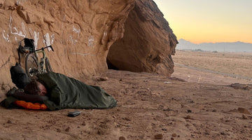 Bikepacker asleep in a bivvy bag under a sandstone overhang at dawn, gravel bike and power bank beside him