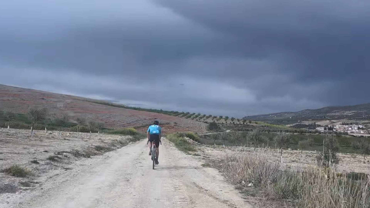 Gravel rider cycling on a dirt path with a stormy sky with olive groves n the distance 