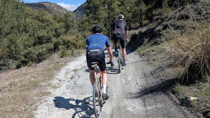 Two gravel cyclists on a dirt trail surrounded by trees and mountains.