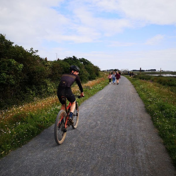 Cyclist on a flat gravel path beside wildflowers, with walkers ahead, on the No Hills to Brighton route.